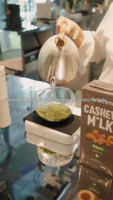 Matcha being whisked in a glass bowl on a scale, with a kettle pouring and an Arummi Barista carton at the right edge.